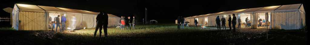 eine Weitwinkelaufnahme des Testfelds bei Nacht. Die Zelte linkt und rechts sind beleuchtet und das Feld liegt im dunkeln.