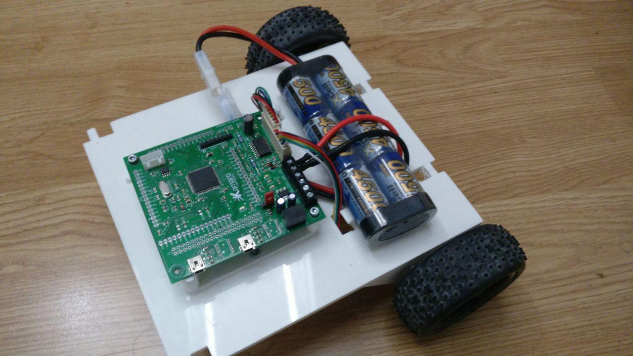 ein kleiner fahrender Roboter, etwa 15 mal 15 cm groß. Angetrieben durch zwei schwarze Räder hinten, gesteuert von einer kleinen grünen Platine, die auf die weiße Grundplatte geschraubt ist. Neben der Platine ist noch ain Akku Pack für die Stromversorgung